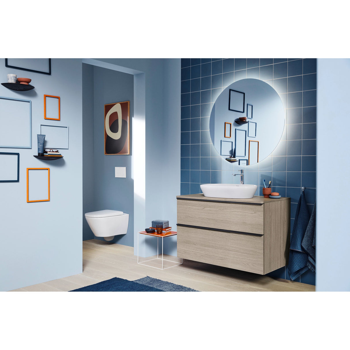 Salle de bain Grohe, meuble vasque bois clair, miroir rond, wc suspendu blanc, déco bleue et orange.