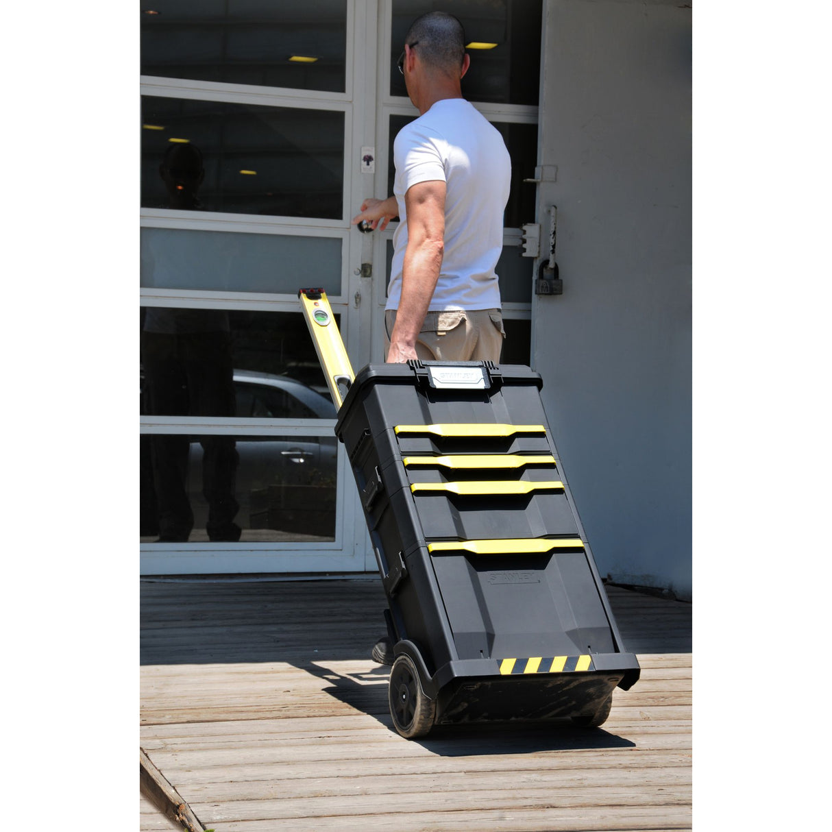 Homme tirant une boîte à outils Stanley noire et jaune, type chariot, devant une porte.
