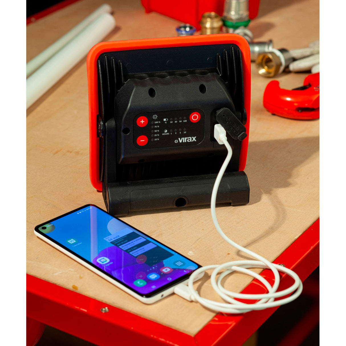 Projecteur LED Virax, rouge et noir, lampe de chantier avec prise USB, posé sur un établi.