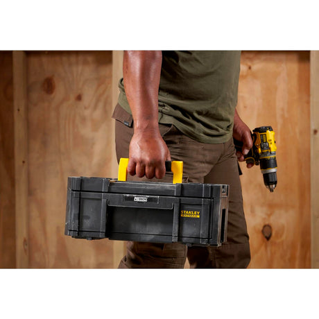 Boîte à outils Stanley Fatmax noire et jaune, transportée à la main, devant un mur en bois.