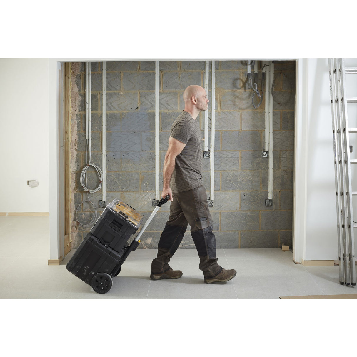 Homme tirant une boîte à outils noire Stanley sur roues, valise de chantier robuste, intérieur en construction.
