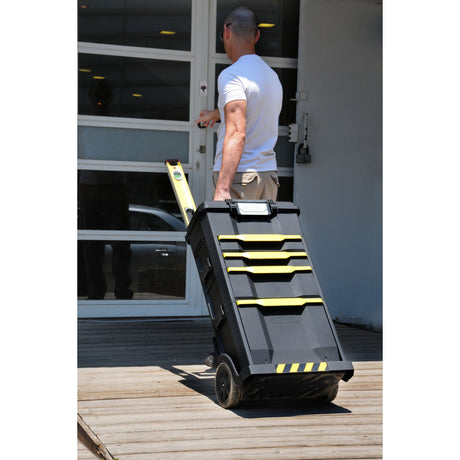 Homme tirant une boîte à outils Stanley noire et jaune, type chariot, devant une porte.