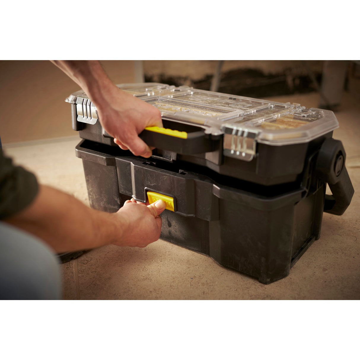 Boîte à outils Stanley noire avec poignée jaune, compartiments transparents, utilisée au sol