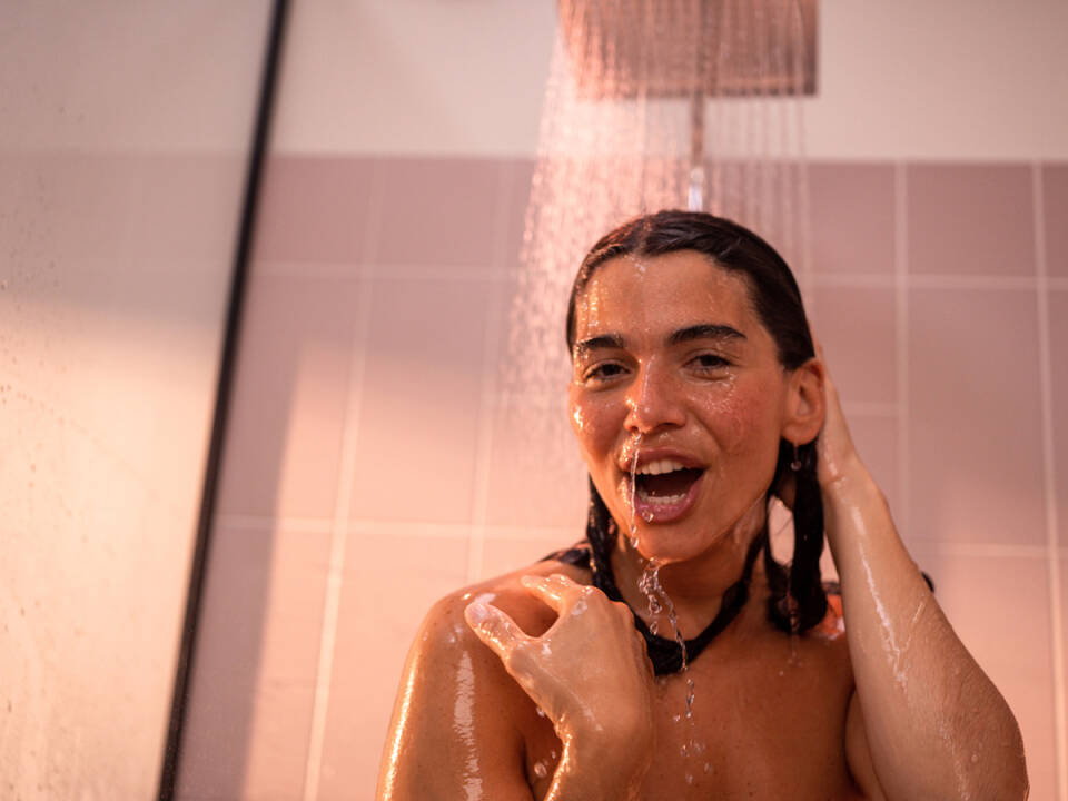 Femme sous douche de tête Hansgrohe Crometta finition chromée dans salle de bain rose.